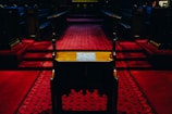 A dimly lit church interior with dark wooden pews and red carpeted flooring, leading towards a slightly elevated pulpit area. The intricate carpet pattern is prominently displayed, and the atmosphere is solemn and reflective.