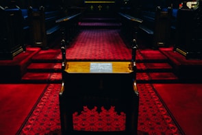A dimly lit church interior with dark wooden pews and red carpeted flooring, leading towards a slightly elevated pulpit area. The intricate carpet pattern is prominently displayed, and the atmosphere is solemn and reflective.