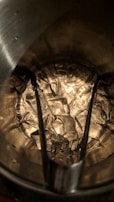 A collection of ice cubes inside a metal bucket with tongs resting on top. The ice appears translucent against the textured surface of the bucket interior, which is reflecting light.