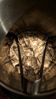 A collection of ice cubes inside a metal bucket with tongs resting on top. The ice appears translucent against the textured surface of the bucket interior, which is reflecting light.
