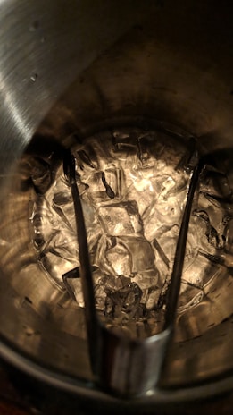 A collection of ice cubes inside a metal bucket with tongs resting on top. The ice appears translucent against the textured surface of the bucket interior, which is reflecting light.