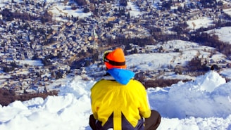 A person dressed in a bright yellow jacket with a blue hood and orange hat sits on a snowy ledge overlooking a sprawling town below. The landscape features snow-covered roofs and fields with trees scattered throughout. The town is surrounded by a snowy and hilly terrain, with clear skies above.