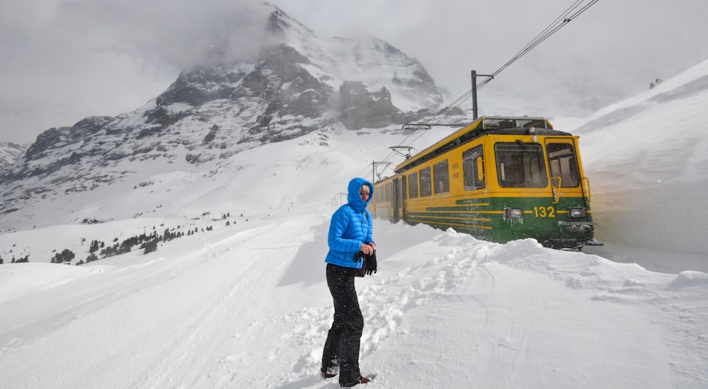 Mann in blauer Jacke und schwarzer Hose steht auf schneebedecktem Boden in der Nähe des gelben Zuges während