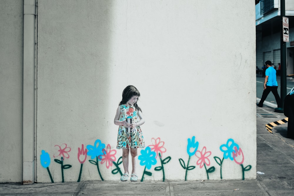 girl in white and pink floral dress standing beside wall with pink flower