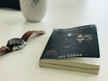 A paperback book with a slightly bent cover lies on a white surface, next to a wristwatch with a brown leather strap and a silver casing. The book cover features an image of two people, and it includes the title and the author's name, Jay Asher. In the background, there is a blurred, white object resembling a vase or container.