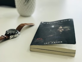 A paperback book with a slightly bent cover lies on a white surface, next to a wristwatch with a brown leather strap and a silver casing. The book cover features an image of two people, and it includes the title and the author's name, Jay Asher. In the background, there is a blurred, white object resembling a vase or container.