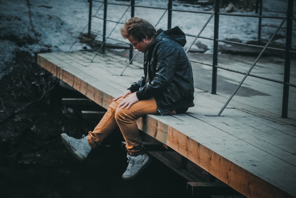 man in black jacket and brown pants sitting on brown wooden bridge during daytime