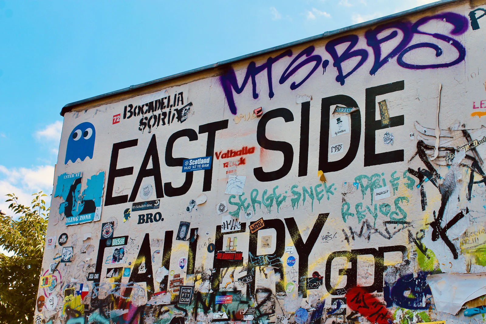 Travel photograph: The words East Side Gallery fill a weathered, sticker-covered section of the Berlin Wall beneath blue sky.