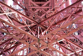 Interwoven metal beams and rivets form a complex lattice structure, showcasing the intricate engineering and architectural design. The beams create an array of intersecting lines and geometric patterns, with light and shadow adding depth and texture.