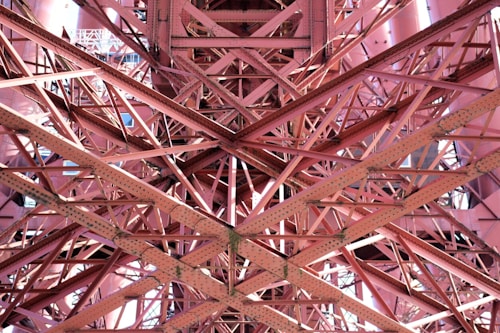 Interwoven metal beams and rivets form a complex lattice structure, showcasing the intricate engineering and architectural design. The beams create an array of intersecting lines and geometric patterns, with light and shadow adding depth and texture.