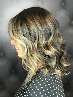 A side profile view of a person with shoulder-length wavy hair, featuring a blend of dark brown and blonde highlights. The background is a tufted black surface, and the person is wearing a polka dot top.