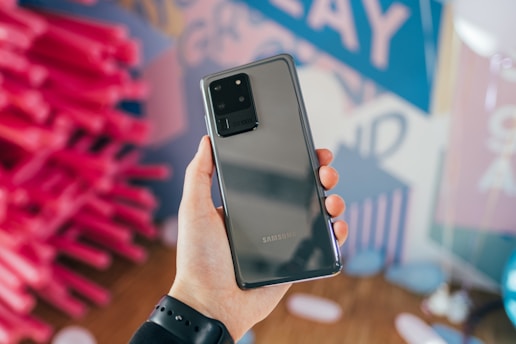 A person holds a Samsung smartphone with a sleek black design and multiple camera lenses on the back. The background is a colorful mix of pink, blue, and purple abstract shapes and blurred text.