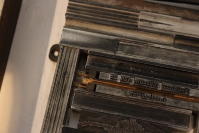 A collection of metal letterpress type forms are arranged in a frame. The letters are reversed and set on a wooden base. There is a decorative floral design etched into one of the metal blocks.