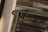 A collection of metal letterpress type forms are arranged in a frame. The letters are reversed and set on a wooden base. There is a decorative floral design etched into one of the metal blocks.