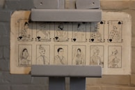 The image features a set of illustrated cards displayed on a stand. The cards depict detailed black-and-white sketches of people in traditional attire, each associated with suits from a deck of cards like clubs and spades. The illustrations convey distinct cultural elements and appear to represent various figures, such as queens, kings, and jacks.