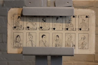 The image features a set of illustrated cards displayed on a stand. The cards depict detailed black-and-white sketches of people in traditional attire, each associated with suits from a deck of cards like clubs and spades. The illustrations convey distinct cultural elements and appear to represent various figures, such as queens, kings, and jacks.
