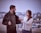 A man and woman are standing on a rooftop engaged in conversation. The man is holding the lapel of his black jacket, and the woman, with long hair, is tugging at his jacket while looking at him with a serious expression. The background features an urban cityscape blurred in the distance.