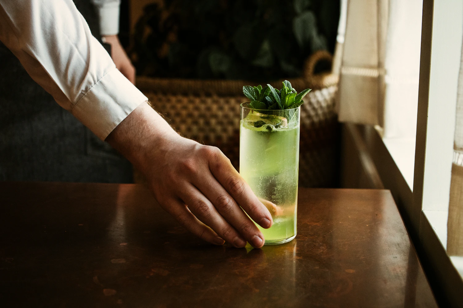 A hand setting down a pale green vermouth and tonic with mint by a bright window