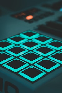 A close-up view of a digital music pad controller featuring twelve square, illuminated buttons arranged in a grid. The buttons emit a teal light, and each button is labeled with a number or letter. The rest of the controller and background are dimly lit, drawing attention to the glowing buttons.