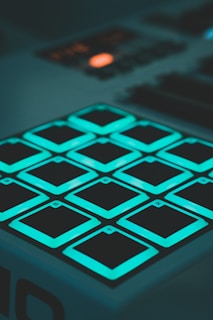 A close-up view of a digital music pad controller featuring twelve square, illuminated buttons arranged in a grid. The buttons emit a teal light, and each button is labeled with a number or letter. The rest of the controller and background are dimly lit, drawing attention to the glowing buttons.