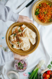 A pair of hands holds a plate with dumplings garnished with herbs and fried onions. Another dish with stir-fried vegetables featuring red peppers and broccoli is visible in the background. The table is set with fresh vegetables, including green onions and tomatoes, and a jar containing sliced onions and herbs.