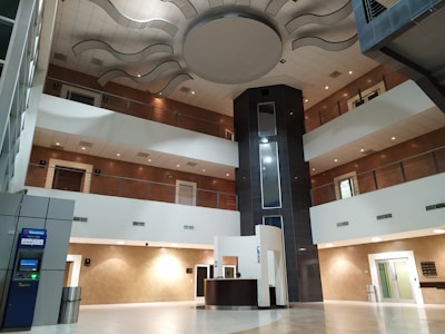 An interior space with a modern design featuring multiple floors and a central atrium. The ceiling has artistic wave-like patterns, and the area includes balconies with metal railings. On the ground floor, there is a reception desk and an ATM to the left. The design incorporates earth tones with white and brown walls, contrasted by a darker vertical column in the center.
