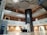 An interior space with a modern design featuring multiple floors and a central atrium. The ceiling has artistic wave-like patterns, and the area includes balconies with metal railings. On the ground floor, there is a reception desk and an ATM to the left. The design incorporates earth tones with white and brown walls, contrasted by a darker vertical column in the center.