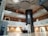 An interior space with a modern design featuring multiple floors and a central atrium. The ceiling has artistic wave-like patterns, and the area includes balconies with metal railings. On the ground floor, there is a reception desk and an ATM to the left. The design incorporates earth tones with white and brown walls, contrasted by a darker vertical column in the center.