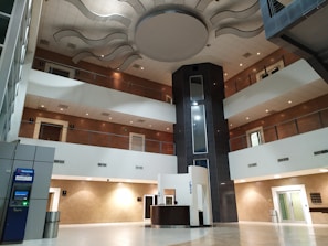 An interior space with a modern design featuring multiple floors and a central atrium. The ceiling has artistic wave-like patterns, and the area includes balconies with metal railings. On the ground floor, there is a reception desk and an ATM to the left. The design incorporates earth tones with white and brown walls, contrasted by a darker vertical column in the center.