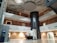 An interior space with a modern design featuring multiple floors and a central atrium. The ceiling has artistic wave-like patterns, and the area includes balconies with metal railings. On the ground floor, there is a reception desk and an ATM to the left. The design incorporates earth tones with white and brown walls, contrasted by a darker vertical column in the center.