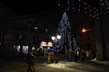 A nighttime scene in a town square featuring a large Christmas tree adorned with blue string lights. People are gathered around, walking and observing the festive decorations. The surrounding buildings are dimly lit, with one displaying the name 'La Locanda' in bright red letters.