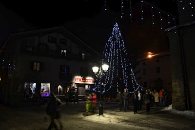 A nighttime scene in a town square featuring a large Christmas tree adorned with blue string lights. People are gathered around, walking and observing the festive decorations. The surrounding buildings are dimly lit, with one displaying the name 'La Locanda' in bright red letters.