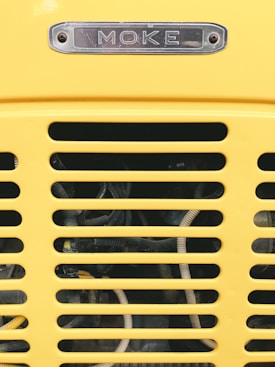 A close-up view of a yellow vehicle grille with horizontal slats, revealing cables and mechanical components behind. Above the grille, a metal nameplate displays the word 'MOKE' in engraved letters.