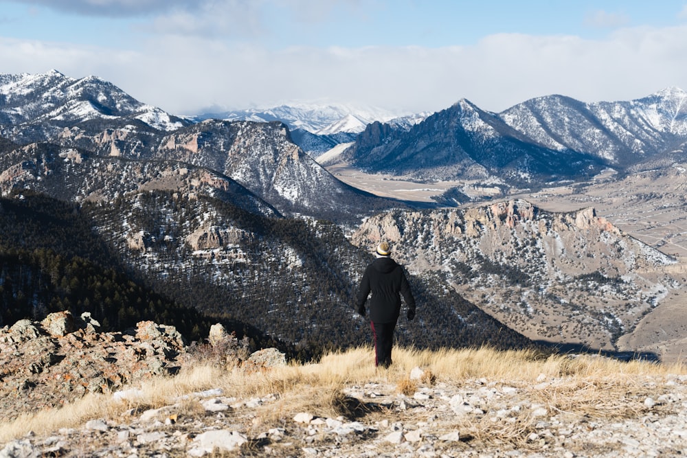 person in black coat standing on brown grass field near gray rocky mountain during daytime