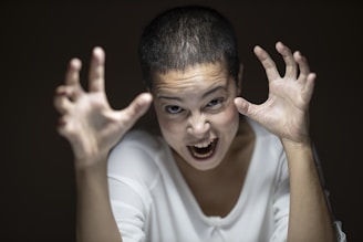A person with short hair is making a dramatic expression, with hands raised and fingers spread wide as if gesturing or roaring. The lighting focuses on the face, highlighting an intense and energetic expression.