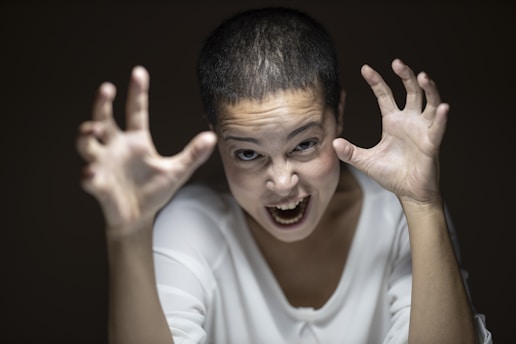 A person with short hair is making a dramatic expression, with hands raised and fingers spread wide as if gesturing or roaring. The lighting focuses on the face, highlighting an intense and energetic expression.