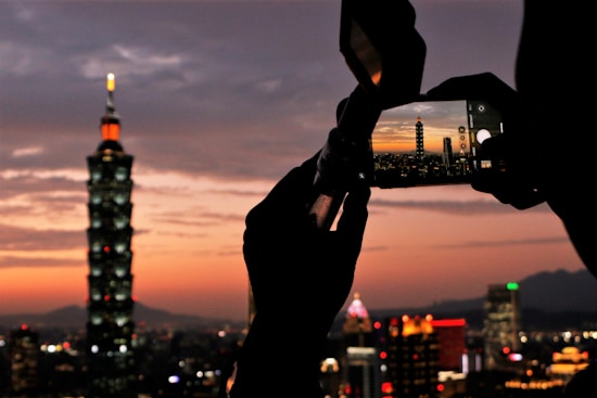 A city skyline at dusk with a prominent skyscraper. A person holds a smartphone, capturing the view. The sky displays shades of orange and purple as the sun sets, creating a picturesque scene.