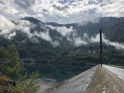 A serene landscape featuring a calm body of water with a clear reflection of the surrounding mountains. Wispy clouds hang low in the sky, partially obscuring the dense forest covering the hills. On the right side, a road with a concrete barrier is visible, leading into the distance. Small houses are nestled among the trees at the base of the hills.