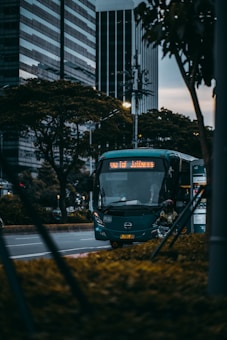 A dark green city bus is driving down a street, bordered by tall office buildings and lush trees. The bus is approaching a bus stop where a person in a helmet and jacket is standing next to a signpost. The scene is set during the early evening, with a moody sky in the background.