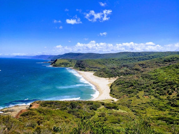 A scenic coastline with a sandy beach bordered by lush green hills and a vibrant blue ocean stretching into the horizon. The sky is clear with scattered clouds, enhancing the bright and sunny atmosphere.