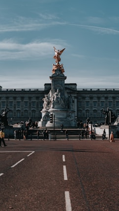 A grand stone monument topped with a golden statue stands prominently in front of an ornate historical building. People are gathered around the monument, suggesting it is a popular tourist attraction. The road leading to the site is wide and relatively empty, and the sky above is clear with scattered clouds.