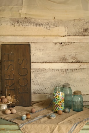 A rustic setting features a wooden sign with the words 'fresh eggs' alongside a collection of jars. One jar is filled with colorful candy-coated chocolates, while other jars are empty and have a vintage look. Brown and blue eggs are arranged on a fabric with blue stripes. The background consists of aged whitewashed wooden planks and sheer fabric drapes down softly.