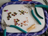 A collection of colorful beads and jewelry-making tools are arranged on a soft surface. Three pairs of pliers with teal handles are scattered around. Various beads, including shiny flower-shaped ones and small square orange beads, are organized in groups. A bunch of golden head pins are also visible.