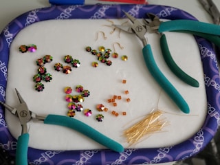 A collection of colorful beads and jewelry-making tools are arranged on a soft surface. Three pairs of pliers with teal handles are scattered around. Various beads, including shiny flower-shaped ones and small square orange beads, are organized in groups. A bunch of golden head pins are also visible.