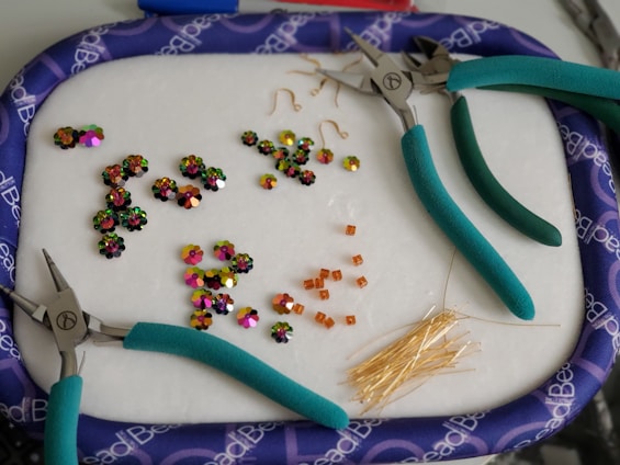 A collection of colorful beads and jewelry-making tools are arranged on a soft surface. Three pairs of pliers with teal handles are scattered around. Various beads, including shiny flower-shaped ones and small square orange beads, are organized in groups. A bunch of golden head pins are also visible.