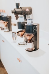 A sleek and modern coffee setup featuring a pour-over coffee maker with copper funnels and clear glass carafes. The coffee equipment is arranged neatly on a minimalist white counter, and steam is visible rising from one of the brewers.