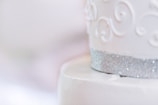 A close-up of an elegantly decorated white cake featuring intricate scroll patterns. The cake is adorned with a shimmering silver band at the bottom, adding a touch of sophistication.