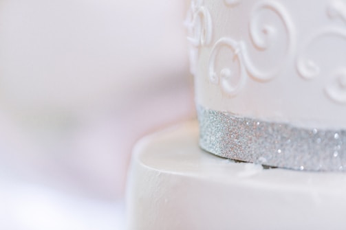 A close-up of an elegantly decorated white cake featuring intricate scroll patterns. The cake is adorned with a shimmering silver band at the bottom, adding a touch of sophistication.