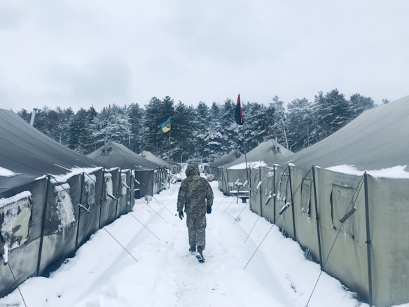 A person dressed in camouflage walks between two rows of large military tents set in a snowy landscape. Snow covers the ground and tents, and two flags are visible in the background. A forest of snow-covered trees lines the horizon.