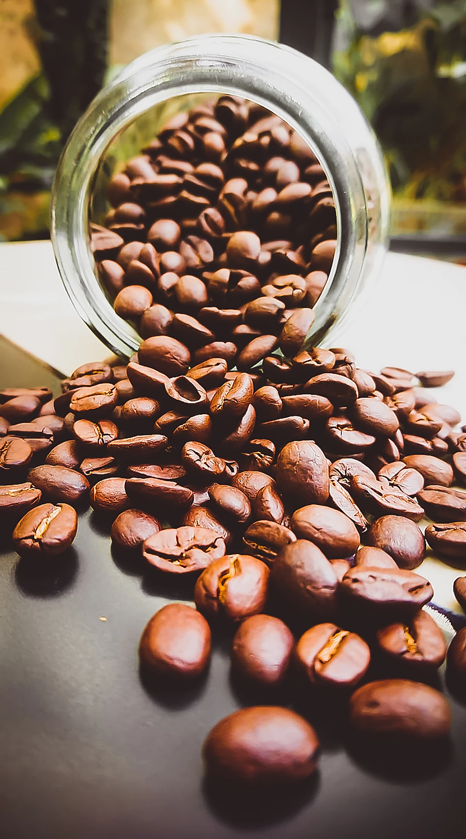 The image shows a glass jar filled with coffee beans spilling out onto a black surface, creating an interesting pattern of brown and white coffee grounds scattered across the table. The jar is positioned on the left side of the frame, while the spilled coffee fills most of the right side of the image.